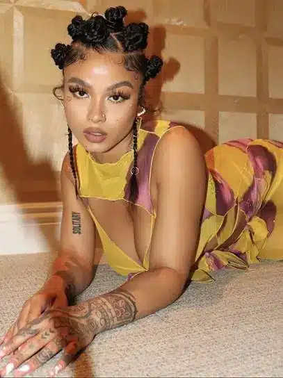 Person with braided hair and tattoos in patterned dress posed on carpeted floor indoors