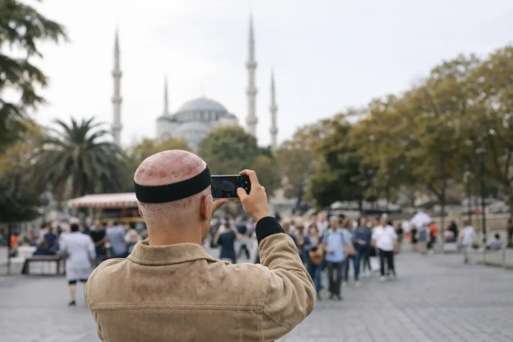 Person taking photo with smartphone in busy outdoor plaza, mosque in background