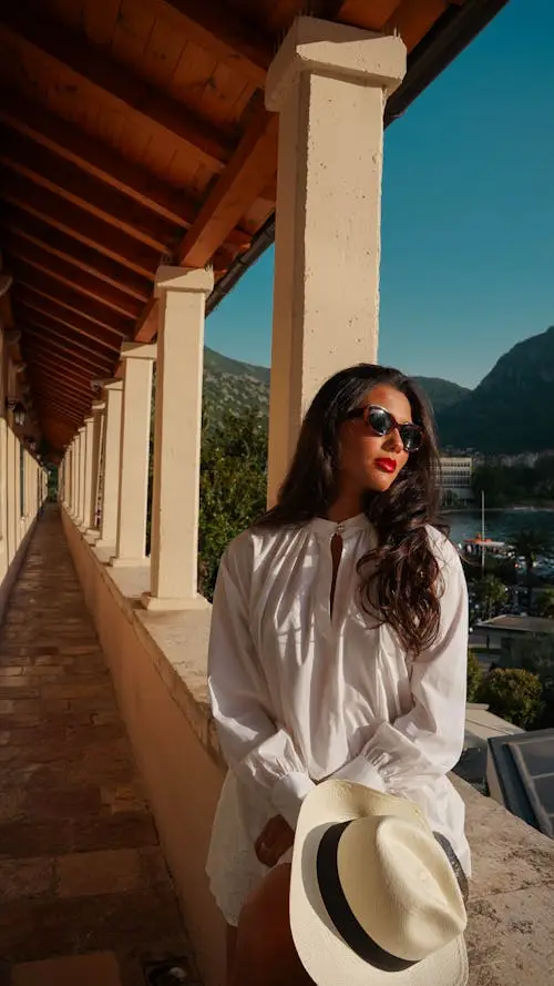 Woman in white dress holding hat, standing on sunlit terrace with mountain view