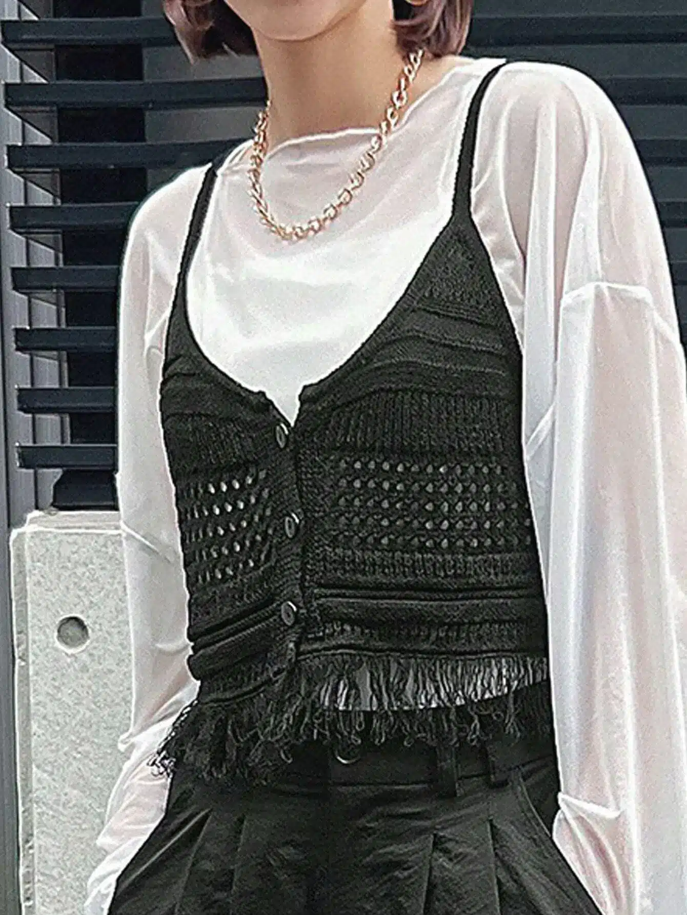 Person wearing black crochet vest over white blouse with gold chain necklace outdoors