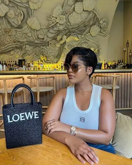 Woman in sunglasses seated at upscale bar with woven Loewe handbag on table