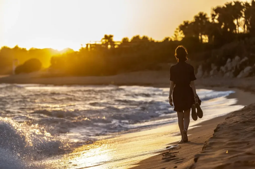 Woman walking barefoot along the shoreline at sunset holding sandals