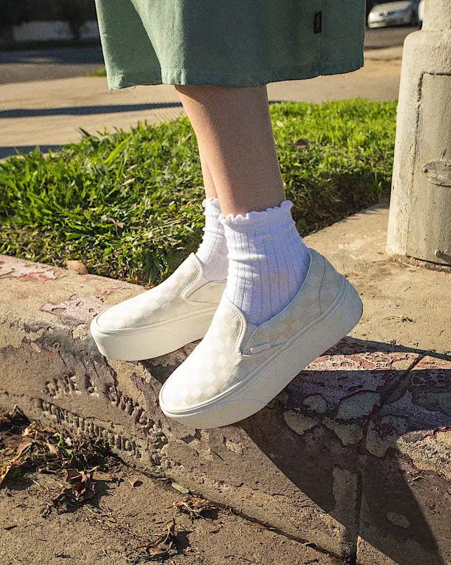 White slip-on sneakers with checkered pattern worn by person in green skirt outdoors