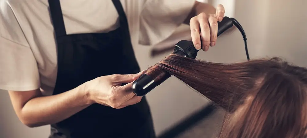 Person styling long brown hair with black curling iron in salon setting