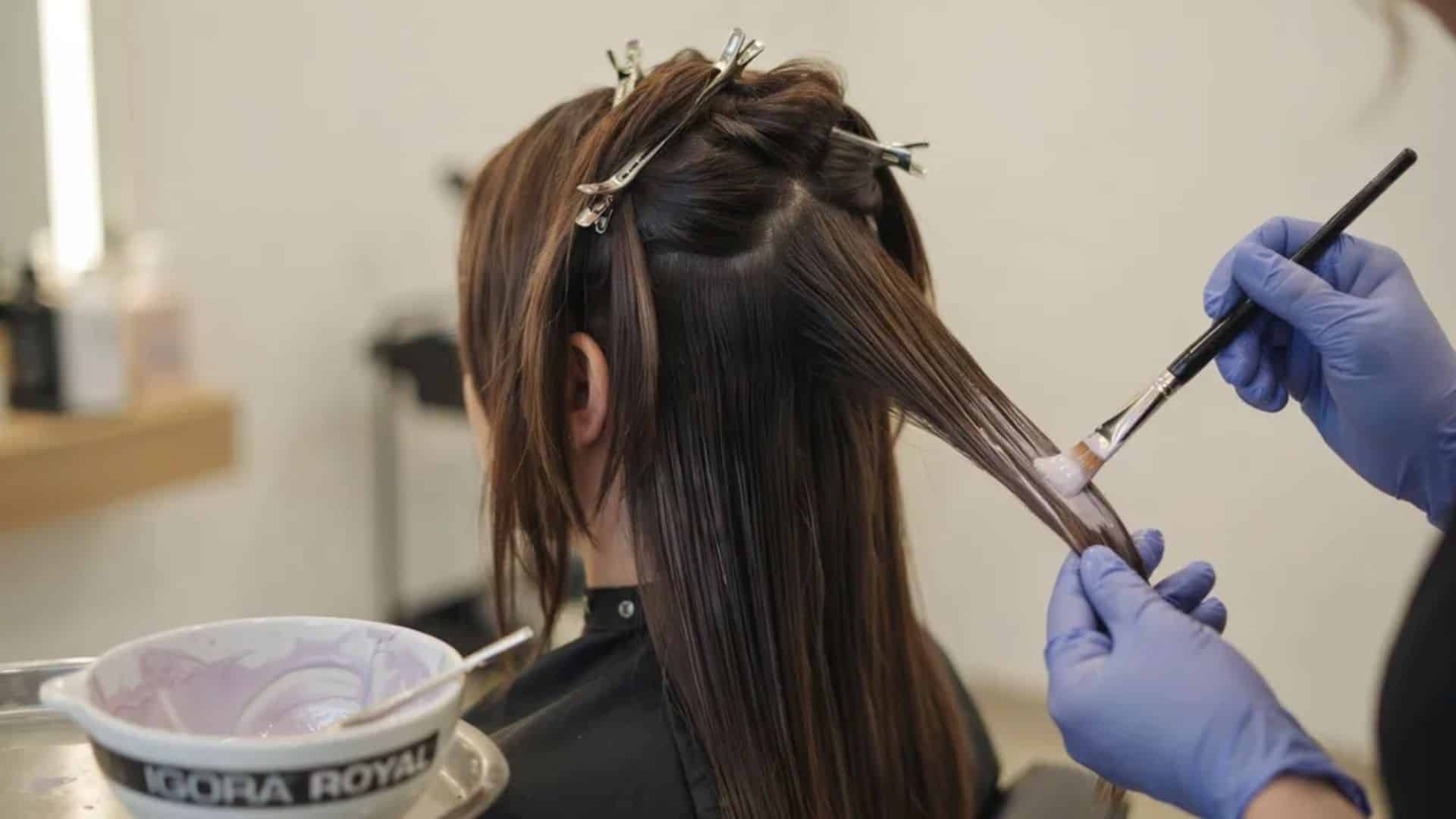 back view of woman applying toner to sectioned dark warm ash-brown hair showing even roots to ends application
