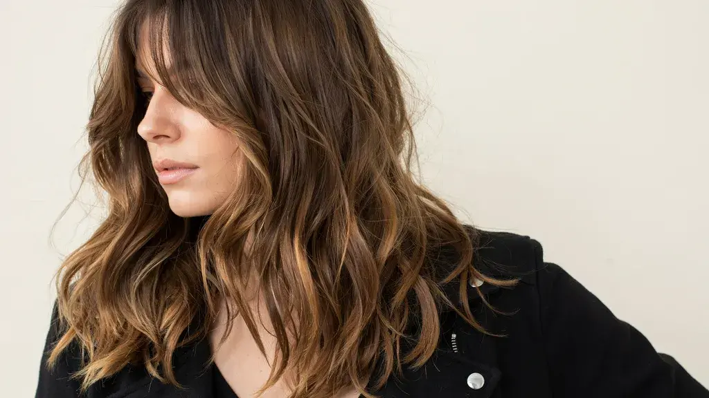 Side profile of a woman with wavy, medium-length brunette hair featuring a subtle shadow root and warm highlights