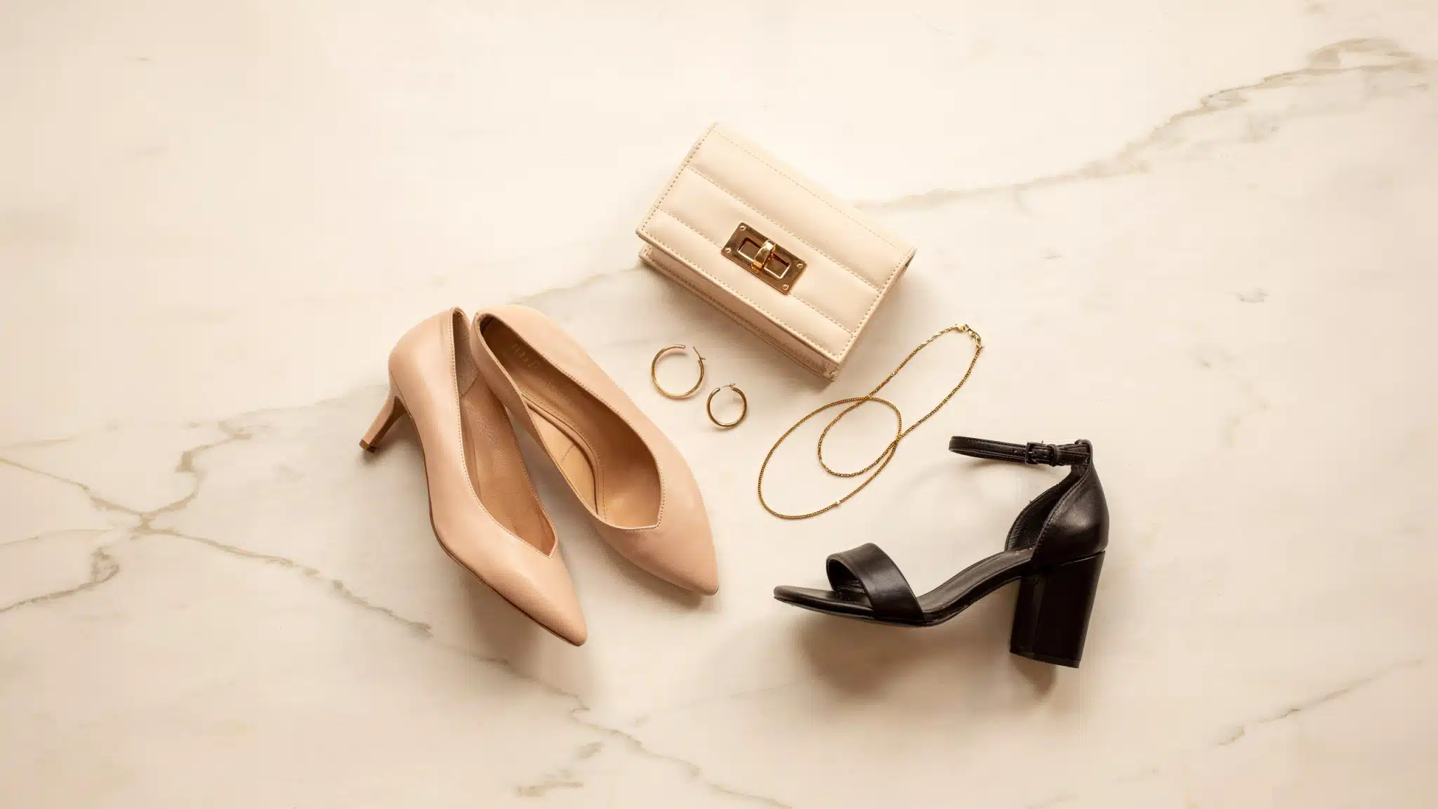 Flat lay of accessories suited to an A-line dress pointed-toe nude heels, a structured ivory clutch, gold hoop earrings, a delicate chain necklace, and a block-heel sandal
