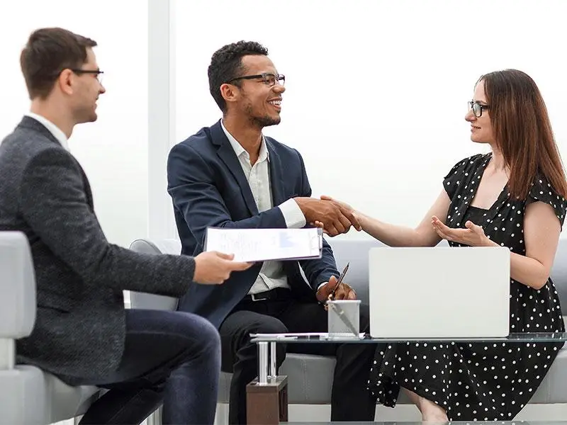 Two professionals shaking hands in modern office, third person holding documents nearby