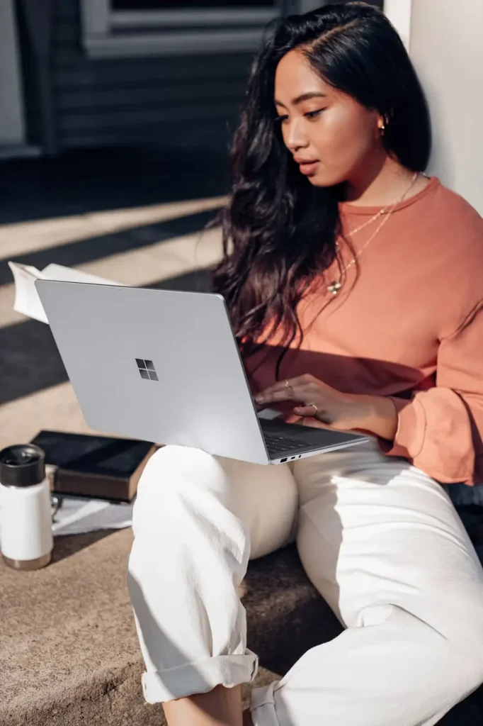Woman in orange sweater working on laptop outdoors with book and coffee mug nearby