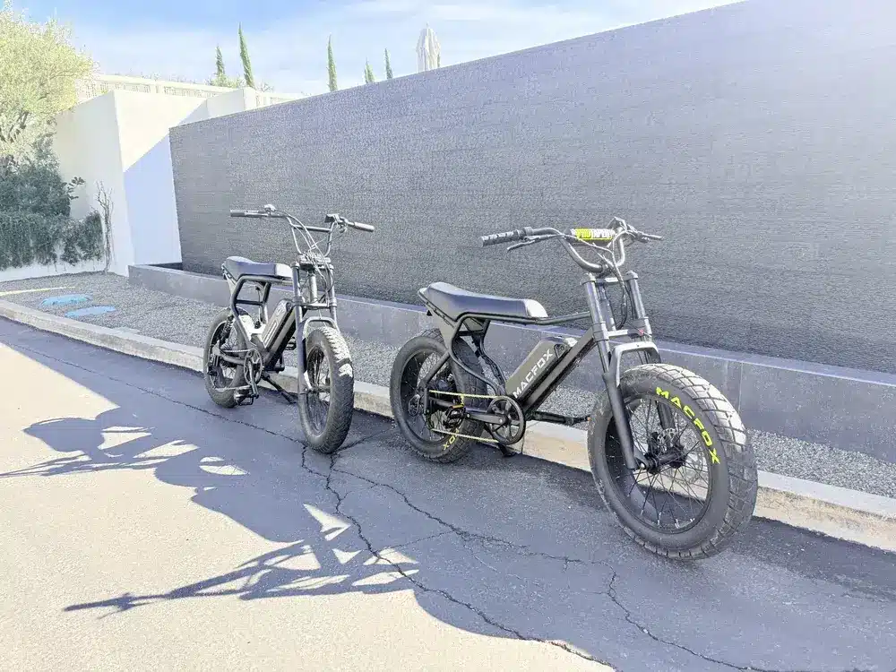 Two black electric bikes parked on a sunlit modern street with a minimalist wall