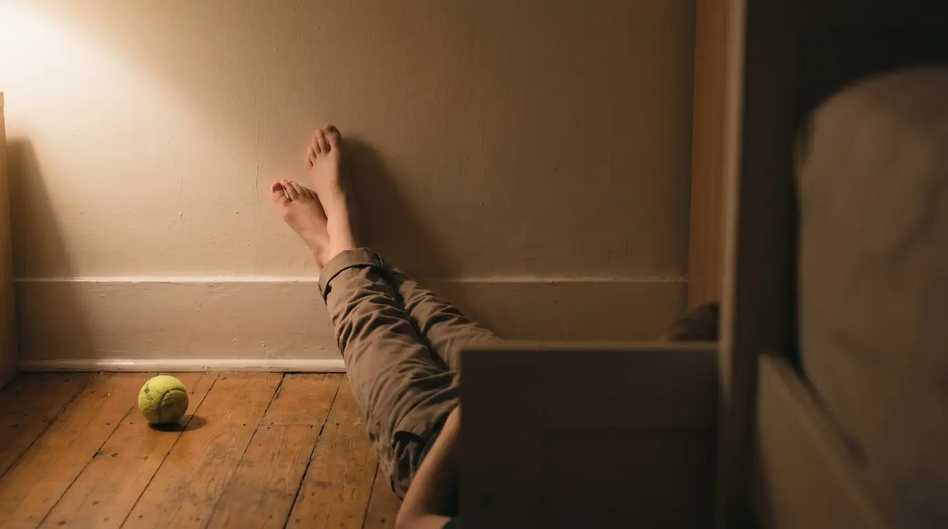 Person lying on wooden floor with legs against wall, tennis ball nearby, warm lighting